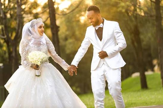 Bride and groom in white wedding attire holding hands in a sunlit garden